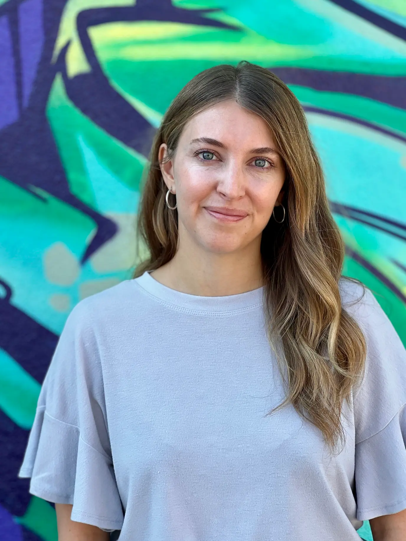 Tessa Brody standing in front of a vibrant, colorful painted wall, wearing a light gray top, with long wavy hair and a warm smile.